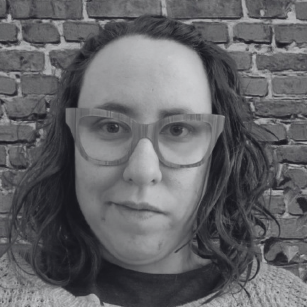 Sadie, a white person with striped glasses and medium-length dark hair, smiles slyly at the camera in a black and white photo. In the background, there is a brick wall. 